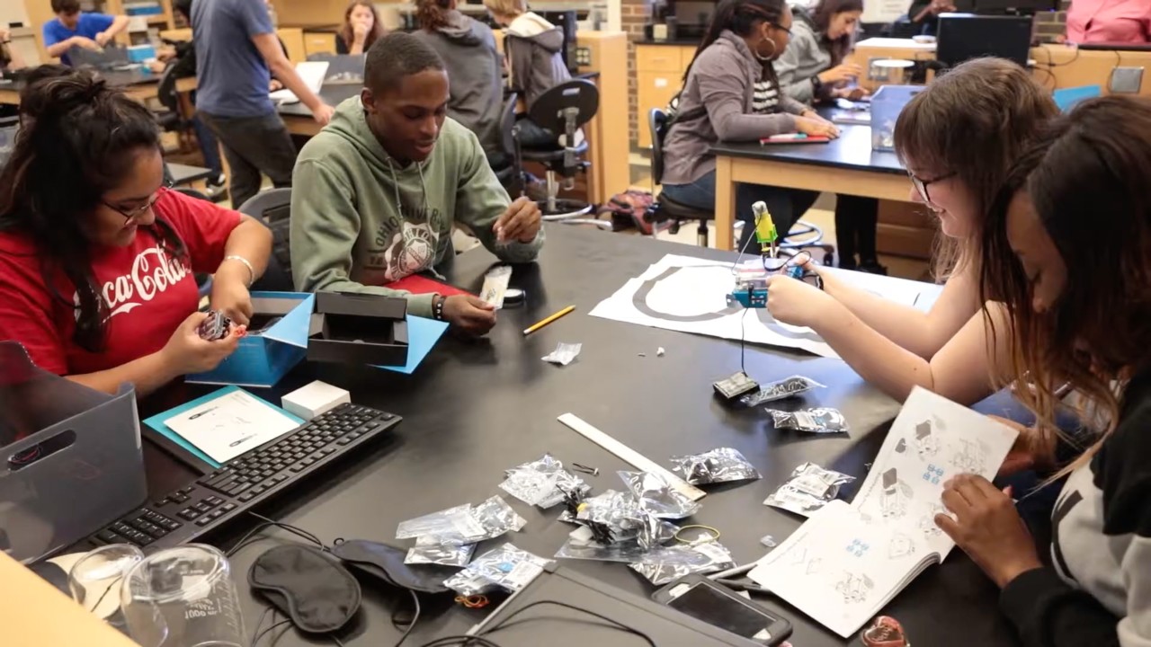 Students sit at a table covered in robotic parts and sensor packages.
