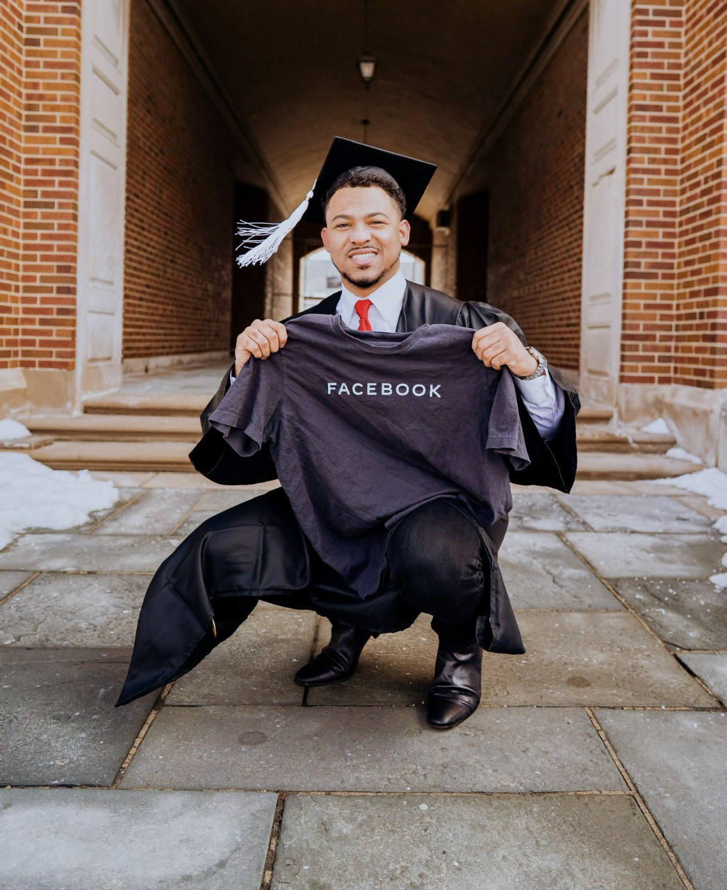 UC's Chandler Rankin wearing a graduation cap and gown kneels in front of McMicken Hall holding a Facebook T-shirt.