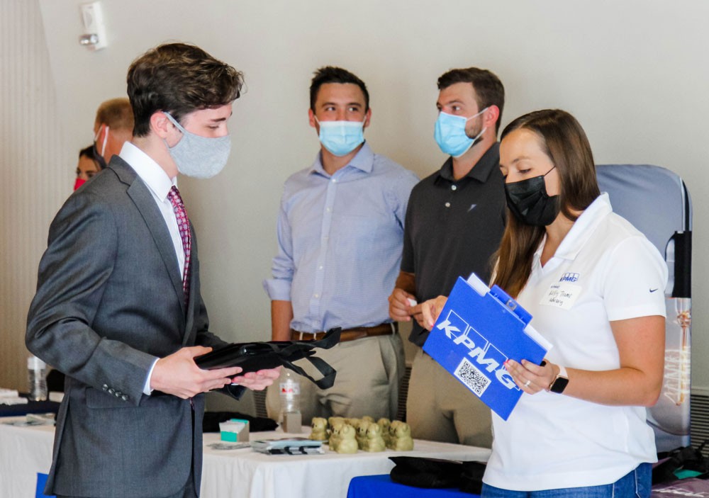 student in grey suit jacket and white shirt and grey mask speaks with a company representative who is wearing a white polo and holding a blue folder with KPMG printed on it. Other company representatives stand in the background