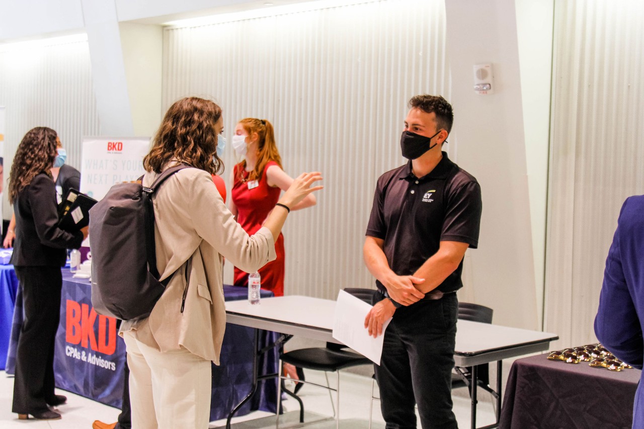 student wearing tan sweater and black backpack stands at an angle speaking to a company representative wearing a black polo and face mask in front of a display table