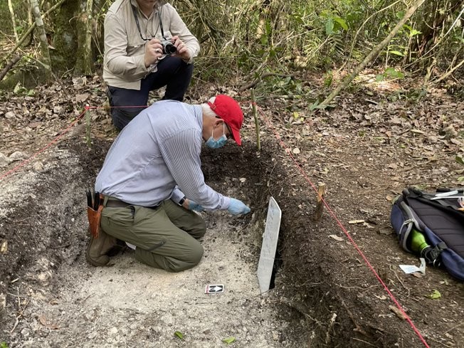 A researcher wearing surgical gloves kneels in an excavation to collect samples for analysis.