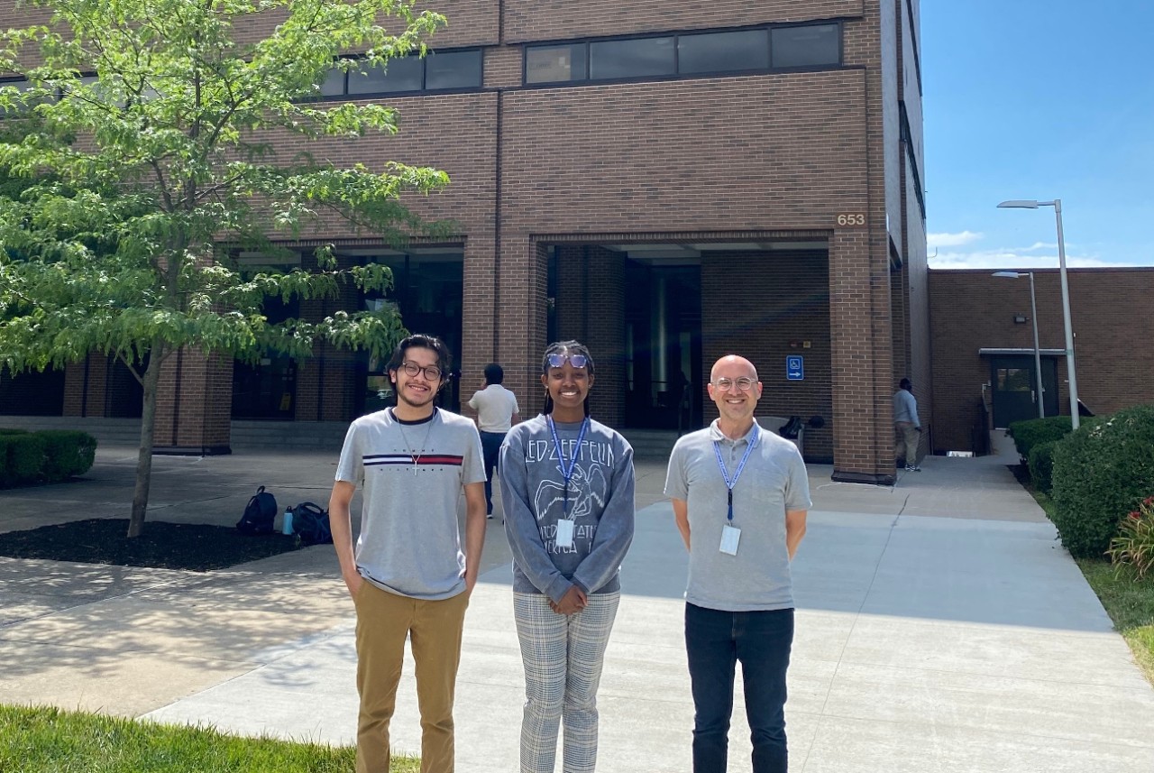 Three people wearing id badges pose in front of a building.