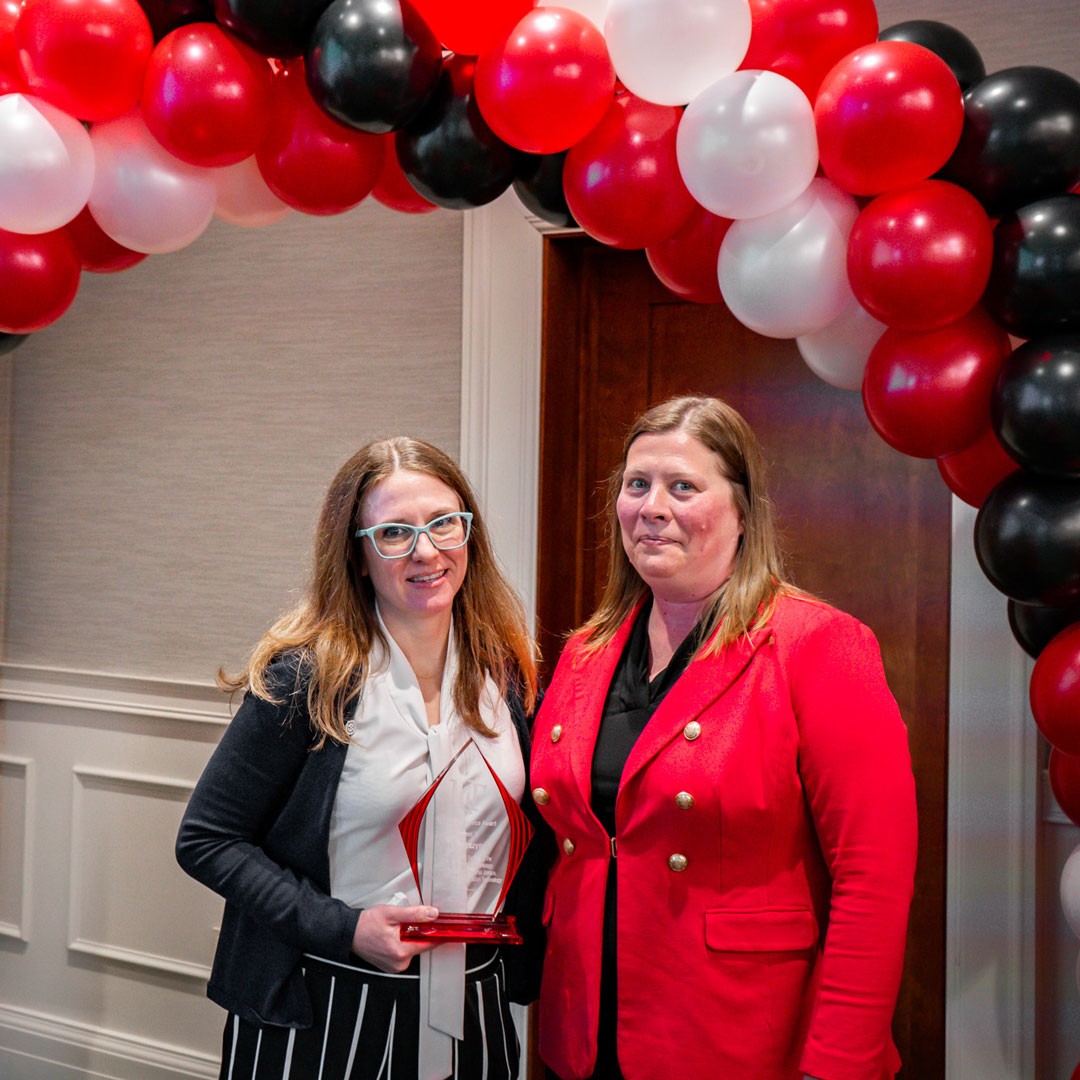two academic professionals stand beneath an arc of red and black balloons