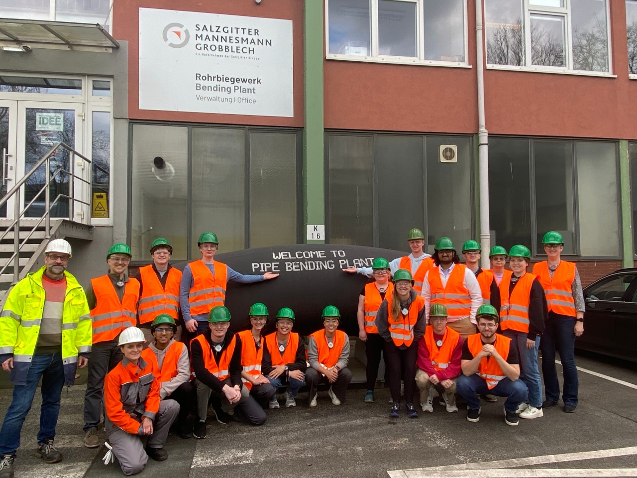 UC group in orange safety vests stands in front of a large bent pipe at pipe-bending plant in Germany