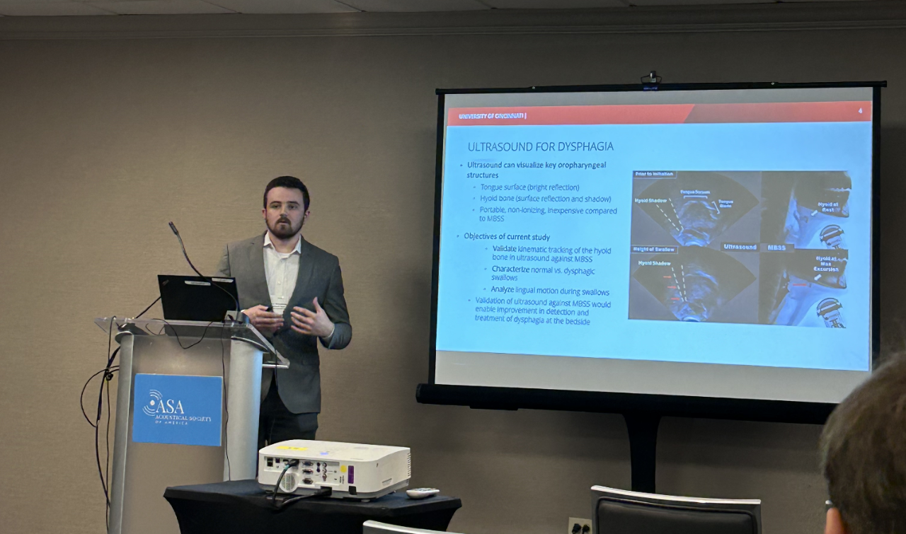 A man in a gray suit stands behind a podium. Next to him is a presentation screen of scientific research.