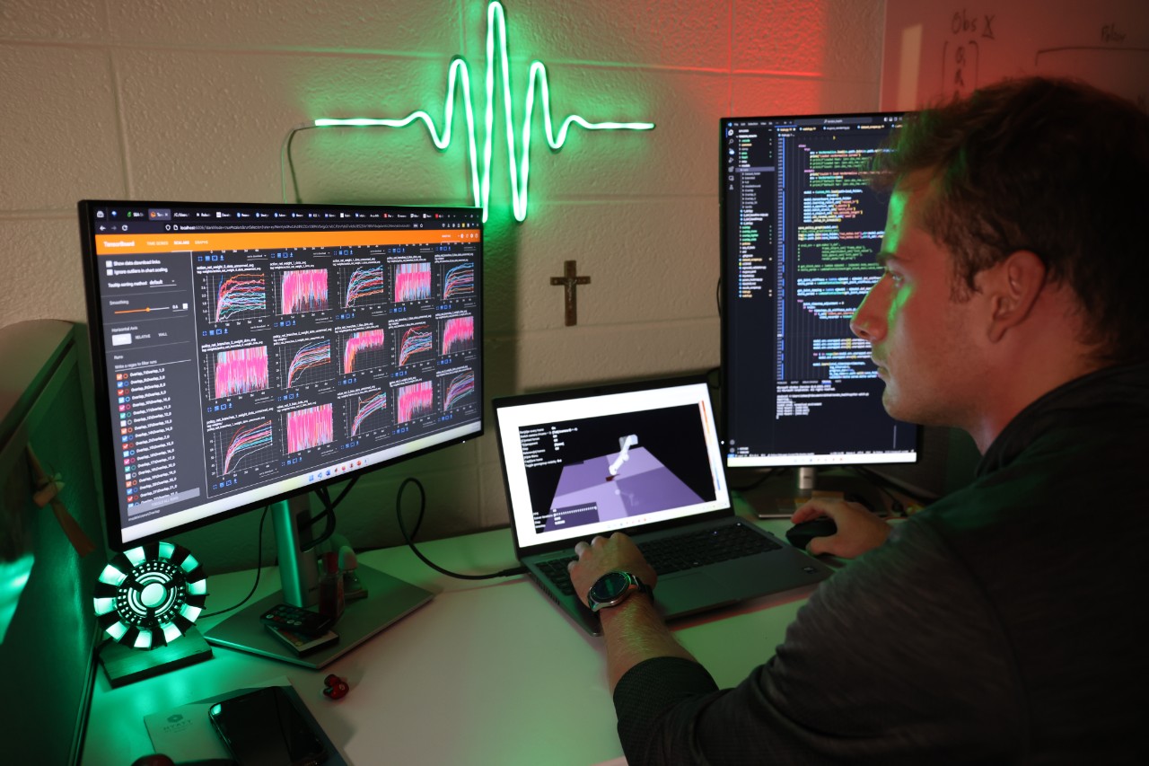 A man sits at a desk with 3 computer screens. The screens show images of graphs and charts.