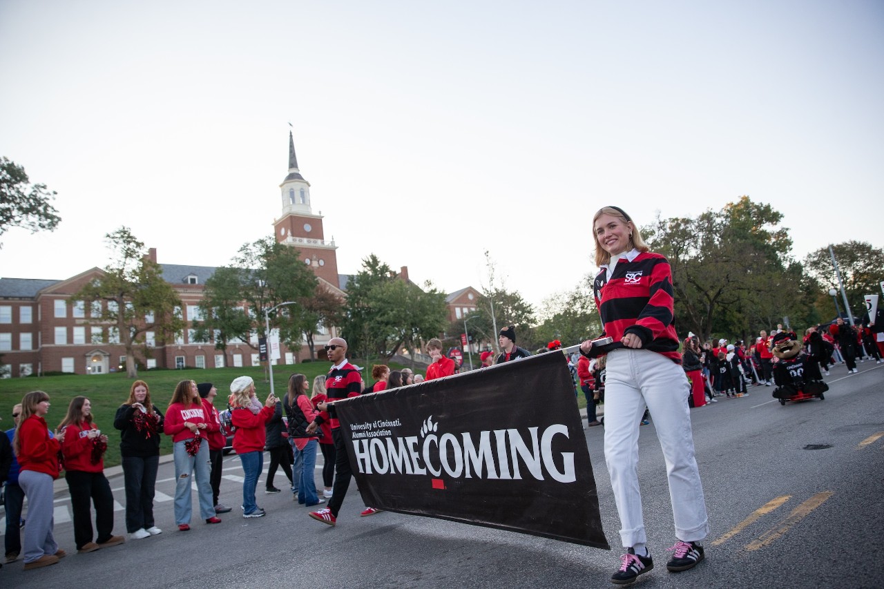 People carrying UC Homecoming banner on street lined with UC fans wearing red and black