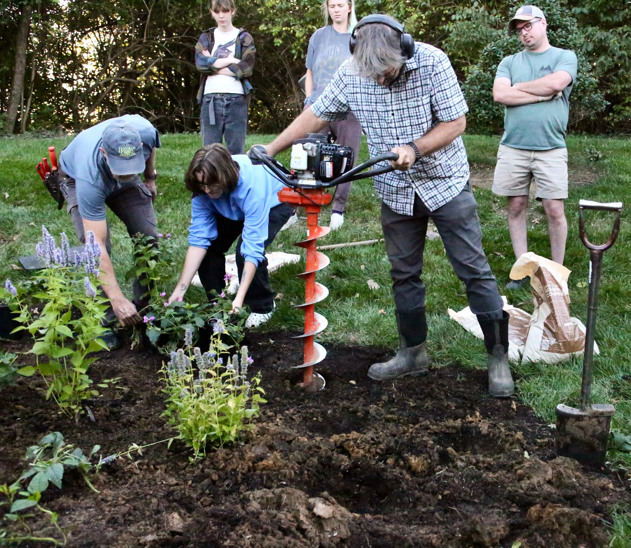 A man in center operates a soil drill as several students observe and help prepare the soil of a new garden.