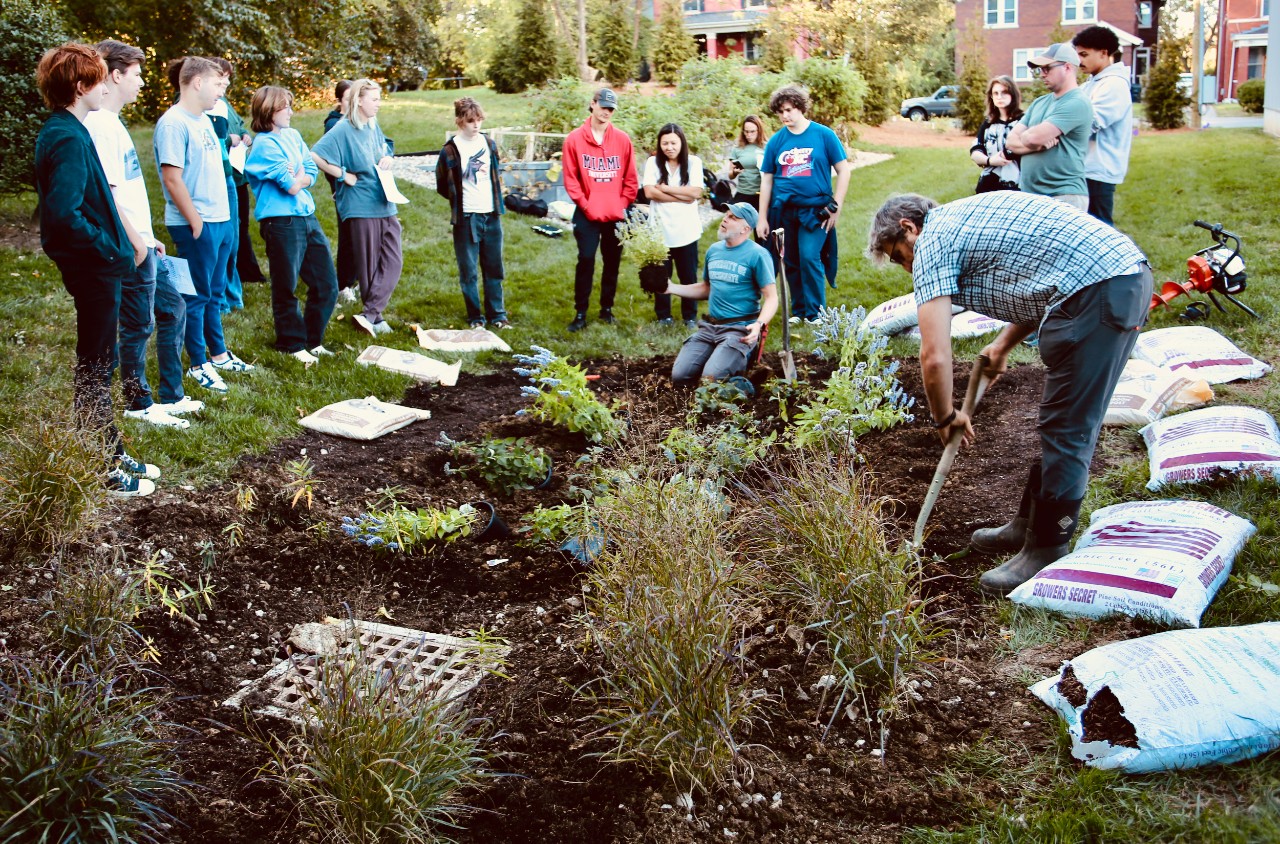 UC Professor Brian Grubb in plaid shirt on right holds a shovel as he digs the soil in a rain garden surrounded by observing students and Professor James Hansel kneeling.