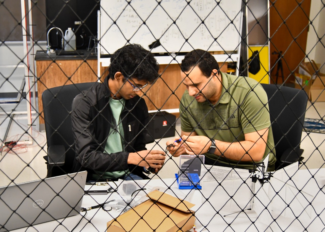 Two students work on a drone's wings behind mesh netting.