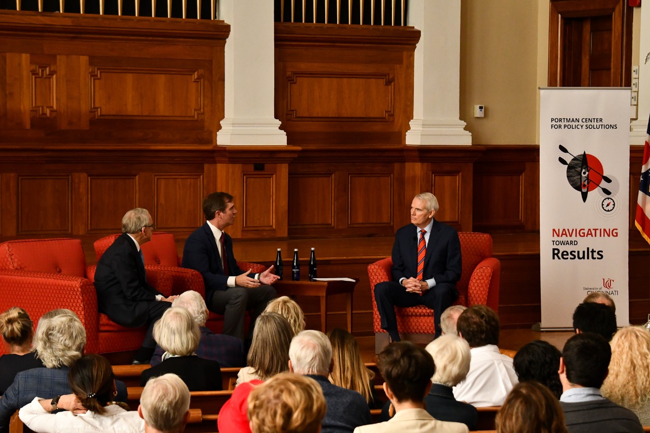 Three speakers seated in chairs on stage talk in front of a large crowd of spectators.