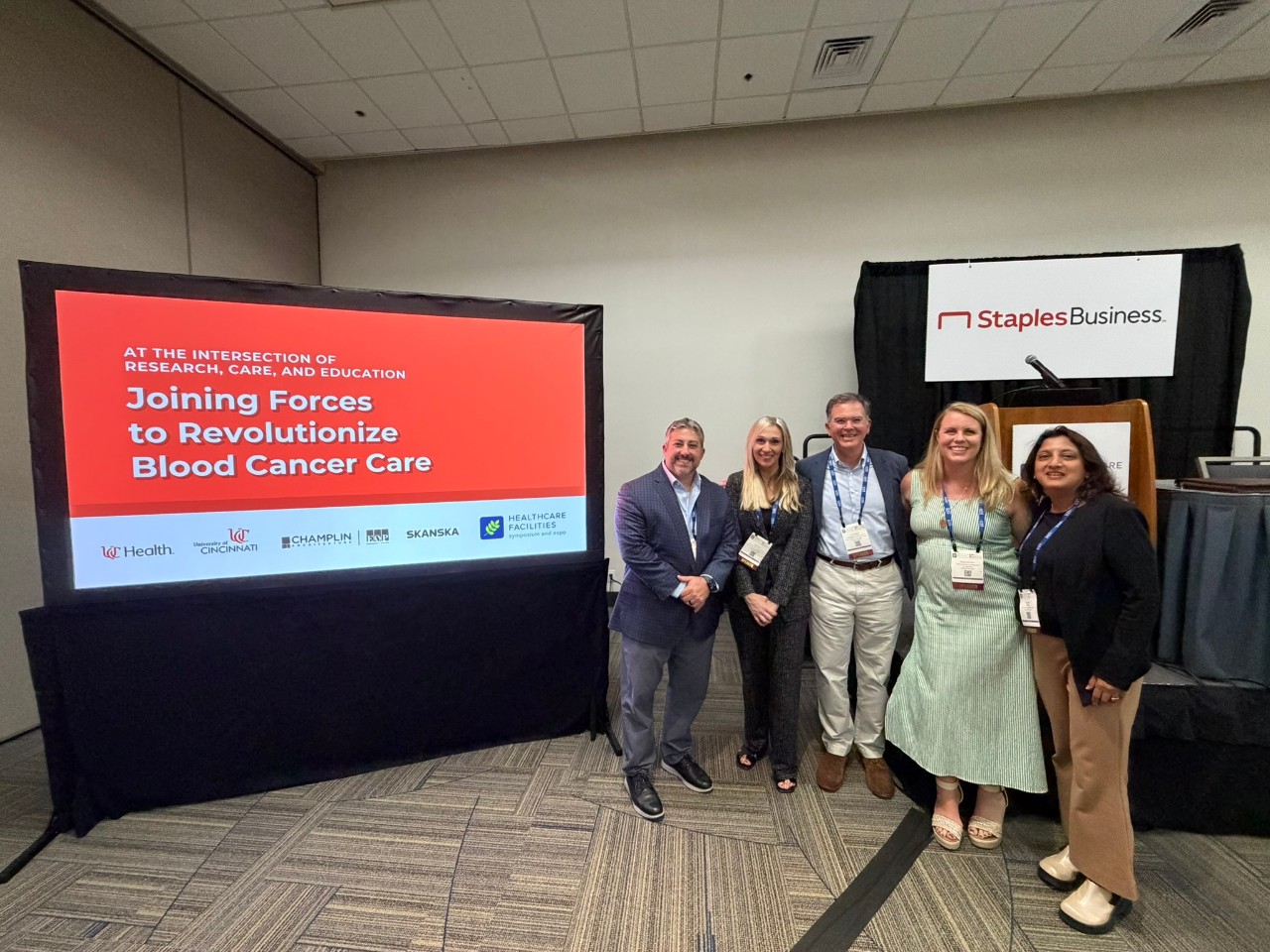 Five people stand together and smile next to a screen displaying their presentation's title